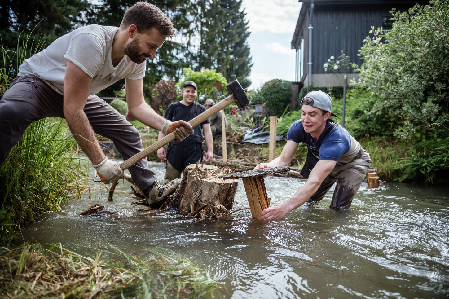 Ein Mann steht im Bach und hält ein rundes Stück Holz. Ein anderer Mann steht am Ufer und hämmert auf das Stück Holz.