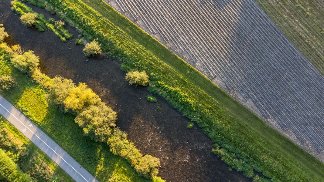 Luftaufnahme eines Bachs, der eng eingezwängt zwischen Verkehrsweg und landwirtschaftlicher Fläche fliesst. 